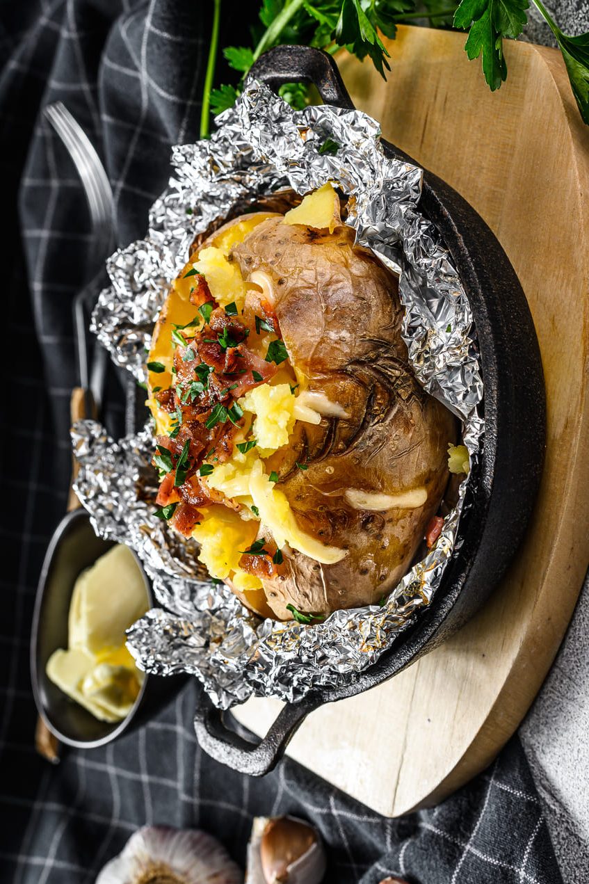 Patata asada con condimentos sobre tabla de madera. Los condimentos son bacon, cebollino y salsa. Está sobre una cazuela con papel de aluminio.