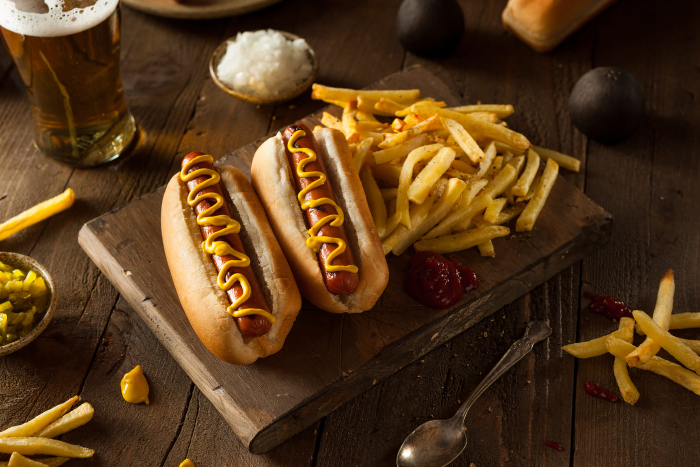 Dos perritos calientes con mostaza, acompañado de patatas y ketchup. Servidos en tabla de madera.