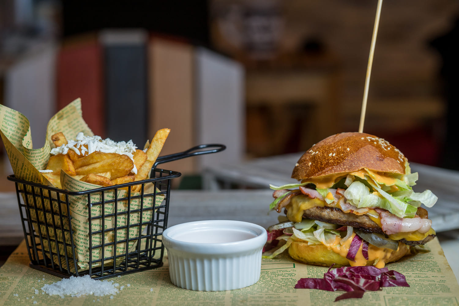 Hamburguesa con patatas en mini freidora y salsa en un cuenco blanco. La hamburguesa es con queso, lechuga, col lombarda y salsa. Tiene un palillo largo clavado arriba.