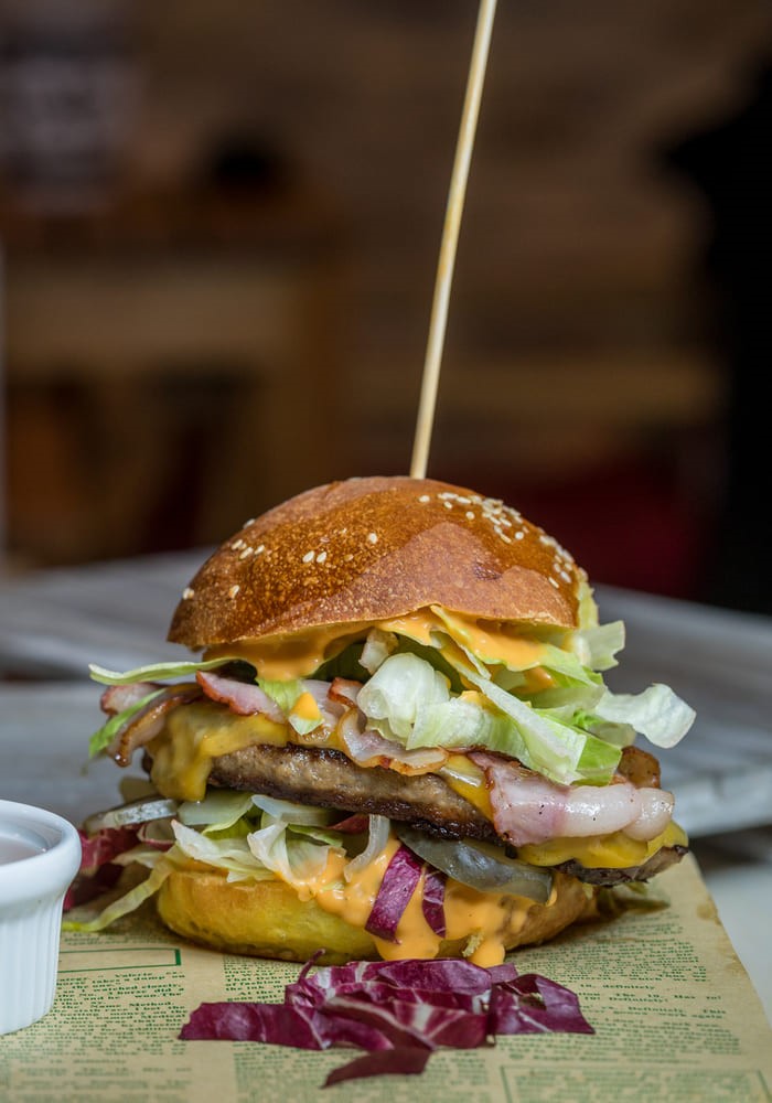 Hamburguesa con queso, lechuga, col lombarda y salsa. Tiene un palillo largo clavado arriba. El fondo está difuminado.