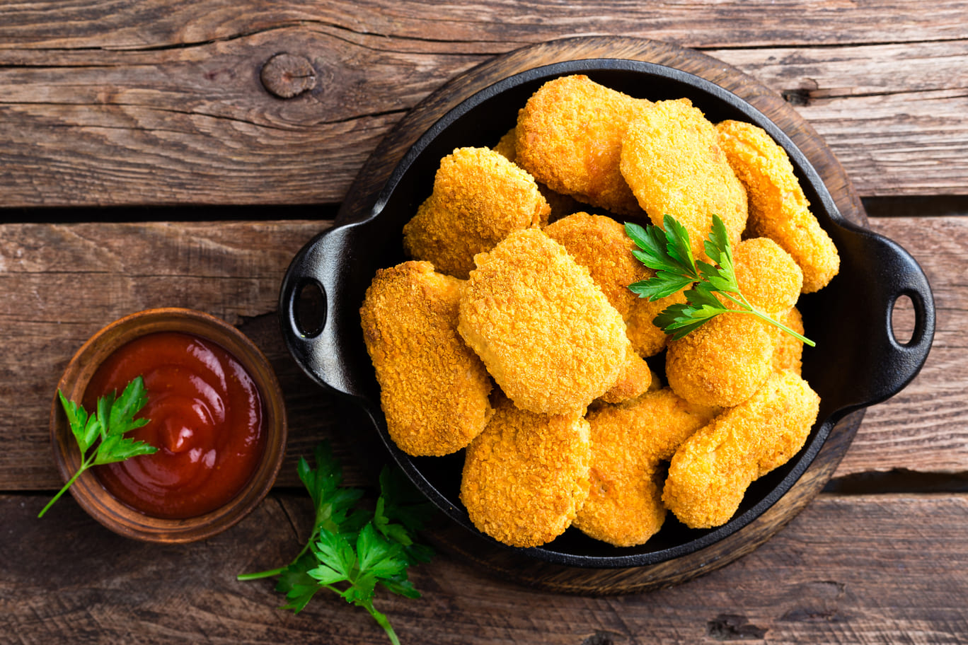 Nuggets de pollo servidos en una cazuela con un cuenco de salsa ketchup aparte. Hay ramas de perejil. El fondo es de madera.